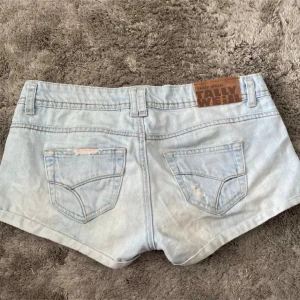 Ljusblå jeansshorts från Tally Weijl - Snygga ljusblå jeansshorts från Tally Weijl med låg midja och klassisk femficksdesign. Shortsen har slitna detaljer framtill och bak, bälteshällor och knappstängning. Perfekta för varma sommardagar och en avslappnad look.