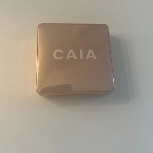 CAIA highlighter i guldig nyans - Snygg highlighter från CAIA i en lyxig, fyrkantig guldig dosa med spegel. Produkten har en skimrande, ljusguldig ton och en vågig, cirkelformad yta som ger glow till ansiktet. Perfekt för att framhäva kindben och ge extra lyster.