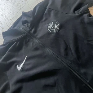Svart PSG Nike tröja - Svart träningsjacka från Nike med PSG-logga på bröstet och vita detaljer på ärmarna. Jackan har hel dragkedja framtill, lång ärm och sportig passform. Perfekt för dig som gillar fotboll och vill ha en clean look. Storlek S, passar någon som är runt 170 nog!