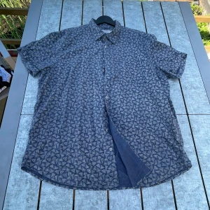 Blå mönstrad skjorta Dressmann 3XL - Snygg blå skjorta från Dressmann i storlek 3XL med vitt triangulärt mönster över hela plagget. Skjortan är tillverkad i en linnemix, har korta ärmar och klassisk krage. Perfekt för dig som gillar en avslappnad men stilren look.