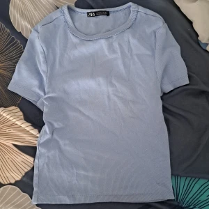 Ljusblå ribbad topp från Zara - En ljusblå ribbad topp från Zara med korta ärmar och rund halsringning med diskret vågig kant. T-shirten är i mjuk bomull och har en enkel, clean look som passar perfekt till jeans eller kjol.