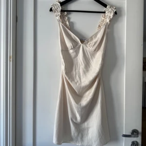 Champagnefärgad kort klänning från Adoore - Superfin kort klänning i champagnefärgad satin från Adoore. Klänningen har breda axelband med volangdetaljer 