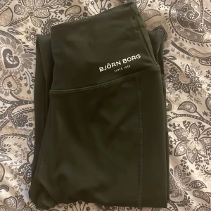 Mörkgröna leggings från Björn Borg - Snygga mörkgröna leggings från Björn Borg med hög midja och tight passform. Tillverkade i stretchigt syntetmaterial som passar perfekt för träning. Diskret logga på höften och coola sömdetaljer längs benen för extra stil.