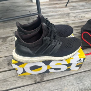 Svarta Adidas Ultraboost sneakers - Svarta Adidas Ultraboost sneakers med stickad ovandel och klassiska tre ränder på sidan. Skorna har en vit, responsiv Boost-sula och svart snörning. Perfekta för dig som vill ha både stil och komfort i vardagen.