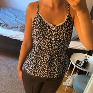 Blommig linnetopp med spets och knappar  - Supersöt marinblå linnetopp med vita små blommor, spetskant vid urringningen och knappar framtill. Toppen har smala axelband och knyt i midjan för en snygg passform. Banden är justerbara🤩