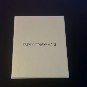 Emporio Armani silverfärgad klocka - Stilren klocka från Emporio Armani med svart urtavla och silverfärgat armband i metall. Klockan har tydliga index, datumvisning och klassisk rund form. Levereras i originalförpackning med Emporio Armani-logga.