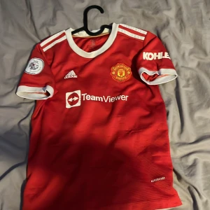 Manchester United Ronaldo 7 Adidas tröja - Röd Manchester United fotbollströja med vita detaljer, officiellt klubbmärke och TeamViewer sponsortryck. Adidas-logga på bröstet och Premier League-märke på ärmen. Namn och nummer Ronaldo 7 på ryggen. Klassisk rund hals och korta ärmar.