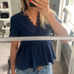 Mörkblå blus med spetsdetaljer - Supersöt mörkblå blus med v-ringning och volang nedtill. Blusen har korta ärmar och fina spetsdetaljer längs halsringningen och ärmsluten. Köpt secondhand 