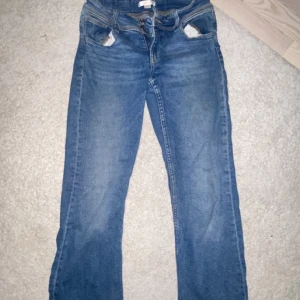 Blå bootcut jeans med fickdetaljer - Säljer ett par klassiska blå jeans med bootcut passform. Jeansen har snygga fickdetaljer med knappar bak och slitningar vid fickorna fram. Perfekta för dig som gillar en tidlös look med lite extra edge.