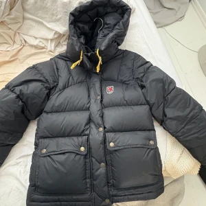 Svart dunjacka från Fjällräven - Säljer en svart dunjacka från Fjällräven med huva och gula snören. Jackan har stora fickor framtill med knappar och klassisk Fjällräven-logga på bröstet. Perfekt för kalla dagar och riktigt snygg passform.