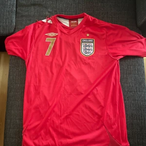 England röd fotbollströja Umbro nr 7 - Säljer en röd England fotbollströja från Umbro med nummer 7 och namnet Carr på ryggen. Tröjan har korta ärmar, guldiga detaljer och broderat landslagsmärke på bröstet. Tillverkad i lätt och ventilerande material, perfekt för match eller träning.