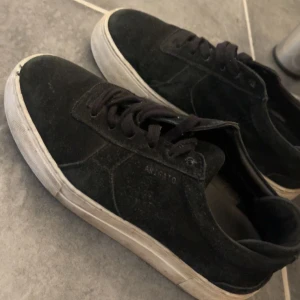 Svarta sneakers från Axel Arigato - Svarta sneakers från Axel Arigato i mocka med vit platt sula och med klassiska snören. Perfekta för dig som gillar stilrena och trendiga skor.