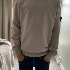 Beige sweatshirt från Stone Island - Säljer en beige sweatshirt från Stone Island med klassisk patch på vänster ärm. Tröjan har rund hals, långa ärmar och ribbade muddar. Tillverkad i mjuk bomull som är skön att bära. Perfekt för dig som gillar stilrena och ikoniska plagg. Är lite sönder vid ärmmudden.