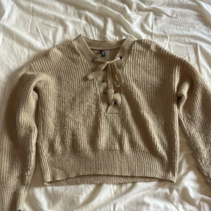 Beige stickad tröja med snörning  - Beige stickad tröja med snygg snörning och metallöljetter vid halsen. Tröjan har lång ärm, ribbade muddar och en lite croppad passform. Perfekt för lager-på-lager och chill höst- eller vinterstil.