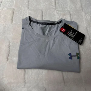 Grå Under Armour t-shirt Threadborne - Snygg grå t-shirt från Under Armour med tryckt logga på bröstet och text längs sidan. Modellen är kortärmad och tillverkad i ett lätt och funktionellt syntetmaterial, perfekt för träning. Rund halsringning och diskreta detaljer på ärmen.