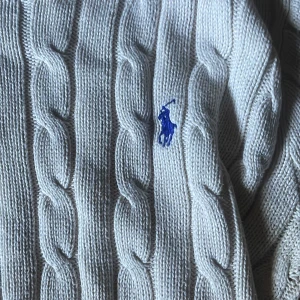 Vit kabelstickad tröja från Ralph Lauren - Vit kabelstickad tröja från Ralph Lauren med klassisk blå broderad logga på bröstet. Tröjan har rund halsringning, långa ärmar och ribbade muddar. Perfekt för dig som gillar preppy och stilren look. Storlek S men passar xs. 