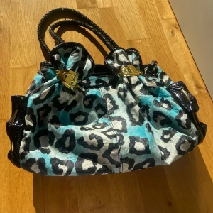 Blå leopardmönstrad axelväska - En vintage axelväska med blått och svart leopardmönster, svarta lackdetaljer och flätade handtag. Väskan har guldiga metalldetaljer, dragkedja och flera fack inuti. En boll från guld detaljerna har lossnat från väskan. 