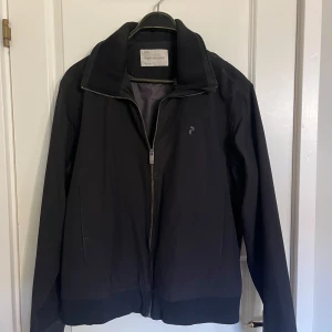 Svart jacka Peak Performance  - Snygg svart jacka från Peak Performance i storlek L. Jackan har dragkedja framtill, hög krage och ribbade muddar vid ärmslut och nederkant. Perfekt för dig som gillar stilren och sportig look. Klassisk logga på bröstet och två fickor på sidorna.