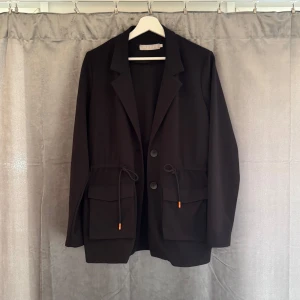 Svart kavaj - Svart kavaj från Pieces med två stora fickor framtill och snygga snörningsdetaljer i midjan som går att justera. Klassisk krage, två knappar och lång ärm. Går över rumpan på mig som är 168.