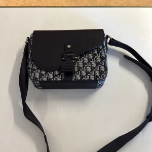 Christian Dior axelväska i skinn och canvas - Snygg axelväska från Christian Dior med klassiskt monogrammönster i blått och vitt. Väskan har lock i svart skinn, justerbar axelrem och coolt spänne framtill. Perfekt storlek för det viktigaste och en riktigt trendig accessoar.