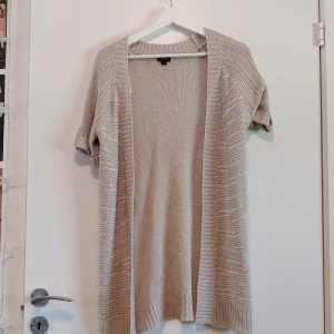 Beige stickad kofta med kort ärm - Beige stickad kofta från LINDEX med korta ärmar och öppen front. Koftan har bred ribbad kant längs öppningen och nederkanten, samt diskreta knappar på ärmarna.