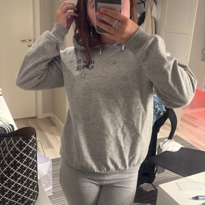 Grå hoodie från Gina Tricot - Mysig grå hoodie från Gina Tricot med texten 'Je m'en Amour' och en liten stjärna framtill. Tröjan har huva, långa ärmar och ribbade muddar. Perfekt för chill dagar och enkel att matcha med jeans eller mjukisbyxor.
