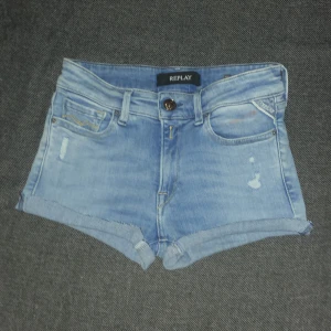 Ljusblå jeansshorts från Replay - Snygga ljusblå jeansshorts från Replay med klassisk femficksdesign, uppvikta benslut och slitna detaljer framtill. Shortsen har låg midja och en normal passform. Perfekta för varma sommardagar och enkel att matcha med allt. Storleken är okänd men passar folk mellan 10-14 år