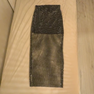 Glittrig långkjol i svart mesh - Säljer en svart långkjol med glittriga stenar över hela tyget. Kjolen är i meshmaterial och har en tight passform som ger en riktigt snygg siluett. Perfekt för dig som vill sticka ut med lite extra bling! Kjolen har en svart underkjol för att inte bli genomskinlig.
