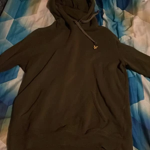 Mörkgrön hoodie från Lyle & Scott - Säljer en mörkgrön hoodie från Lyle & Scott med klassisk gul logga på bröstet. Tröjan har huva med snörning och en stor magficka. Perfekt för dig som gillar en enkel och stilren look.
