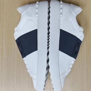 Valentino Garavani Open sneakers vit/blå - Snygga Valentino Garavani Open sneakers i vitt skinn med bred mörkblå rem över mitten. Skorna har ikoniska nitar på hälen och längs sulan, samt vita snören. Perfekt för dig som vill ha en clean men edgy look.