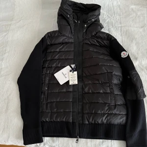 Svart Moncler pufferjacka med huva (cardigan) - Svart pufferjacka från Moncler med huva och ribbade ärmar. Jackan har dragkedja framtill, två fickor och ikonisk Moncler-logga på ärmen. Materialmix av dun, polyester och stickade detaljer ger en clean och sportig vibe.