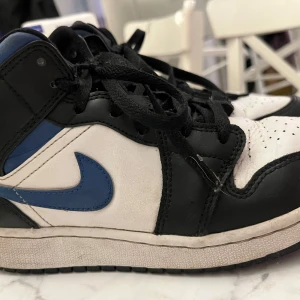 Nike Air Jordan 1 svart/blå sneakers - Säljer nu mina fina Snygga Nike Air Jordan 1 sneakers i svart, vitt och blått skinn. Eftersom det är inte riktigt min stil längre ,Klassisk high-top modell med svart snörning och perforerad tå. Ikonisk blå swoosh och Jordan Wings-logga på sidan. Perfekta för dig som vill sticka ut med streetstyle.