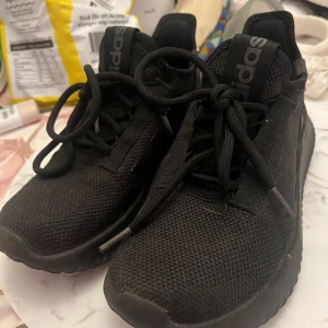 Svarta Adidas sneakers med mesh - Snygga helsvarta Adidas sneakers med klassiska tre ränder på sidan. Ovandelen är i mesh för bra andningsförmåga och skon har en tjock, bekväm sula. Perfekta för dig som gillar en clean och sportig look. Skorna har snörning och rund tå.