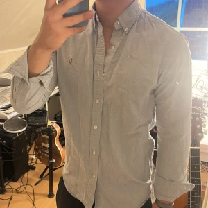 Ljusgrå skjorta från Polo Ralph Lauren - Säljer en stilren ljusgrå skjorta från Polo Ralph Lauren med klassisk button-down krage och broderad logga på bröstet. Skjortan är i bomull, har lång ärm och knäppning framtill. Perfekt för dig som gillar en clean och tidlös look.