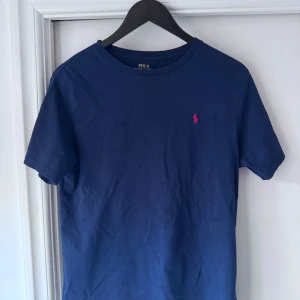 Marinblå t-shirt från Polo Ralph Lauren - Snygg marinblå t-shirt från Polo Ralph Lauren med klassisk passform. T-shirten har korta ärmar och är gjord i mjuk bomull. På bröstet finns den ikoniska rosa Polo-loggan broderad. Perfekt för en clean och stilren look.