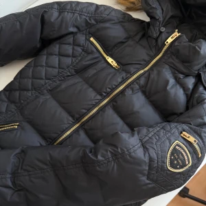 Svart Rockandblue dunjacka med fuskpäls - Snygg svart dunjacka från Rockandblue med guldiga dragkedjor och detaljer. Jackan har quiltad design, flera fickor och en stor huva med fluffig fuskpäls. Perfekt för kalla dagar och ger en cool streetstil. Passformen är bra och jackan är långärmad. Köpte den 2 år sedan och använde den bara en vinter men den har gått sönder lite längst ner därav priset men förutom de finns d ringa skador. Har tagit av pälsen men skickar den med jackan. Ord pris 3699kr, pris kan diskuteras.