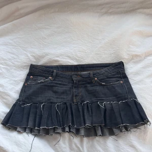 Mörk jeanskjol med volang från Weekday - Säljer en mörkblå jeanskjol från Weekday med volangkant och råa, fransiga kanter. Kjolen har klassiska jeansdetaljer som fickor fram och bak samt bälteshällor. Perfekt för dig som gillar edgy och trendig stil.