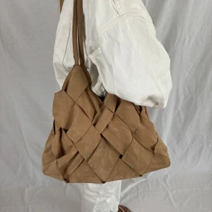 Beige axelväska i mocka med flätad design - Säljer en stor och mjuk axelväska i beige mocka med unik flätad look. Väskan har långa handtag som gör den enkel att slänga över axeln och rymmer allt du behöver till vardags. Perfekt för dig som gillar en chill och trendig stil.