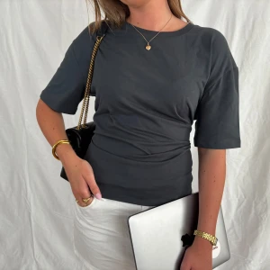 Mörkgrå t-shirt med knyt i ryggen - Snygg mörkgrå t-shirt i bomull med korta ärmar och rund hals. T-shirten har en unik detalj med knytning i ryggen som ger en cool och trendig look. Perfekt att styla med jeans eller kjol för en avslappnad vibe.