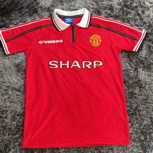 Retro Manchester United tröja  - Klassisk Manchester United fotbollströja från Umbro i rött med vita och svarta detaljer. Kortärmad modell med vit krage och svarta ränder på axlarna. Broderat klubbmärke och SHARP-tryck på bröstet. Använd minst en gång.