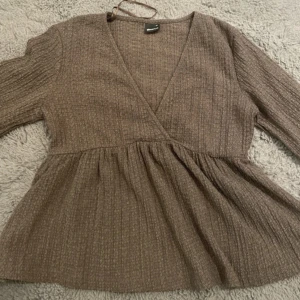 Brun omlottopp med volangärm - Supersöt brun omlottopp från gina tricot med v-ringning och långa volangärmar. Toppen har en ribbad struktur och knytband i nacken. Perfekt för dig som gillar en avslappnad men ändå trendig look.