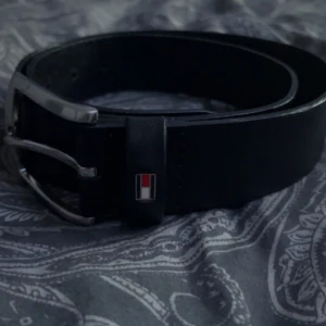 Svart skärp från Tommy Hilfiger - Snyggt svart bälte från Tommy Hilfiger i skinn med klassiskt silverfärgat spänne. Detalj med Hilfigers logga i rött, vitt och blått vid öglan. Perfekt accessoar för att lyfta din outfit.
