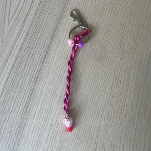 Rosa flätat nyckelband med milkshake-berlock - Sött handgjort nyckelband i rosa flätat snöre med små pärlor i rosa och lila. Längst ner hänger en miniberlock formad som en milkshake i rosa och vitt. Perfekt att hänga på väskan eller nyckelknippan för en gullig touch.