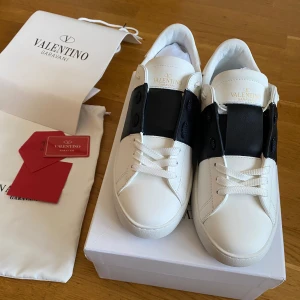 Valentino Garavani Open sneakers vit/svart - Snygga Valentino Garavani Open sneakers i vitt skinn med bred svart läderdetalj över vristen. Klassisk rund tå, vita snören och diskret logga på plösen. Sulan har coolt mönster och nitar baktill för extra edge.