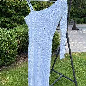 Vit stickad långklänning Madlady - Supersnygg vit stickad långklänning från Madlady med ett unikt mönster, lång ärm på ena sidan och dubbla smala axelband på andra. Asymmetrisk offshoulder-design som ger en cool och trendig vibe. Perfekt för sommarens alla tillfällen!