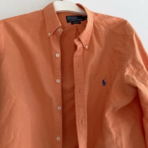 Orange skjorta Polo Ralph Lauren M - Snygg orange skjorta från Polo Ralph Lauren i slim fit-modell. Skjortan har klassisk krage, knäppning framtill och det ikoniska blå broderade logot på bröstet. Tillverkad i mjuk bomull och har långa ärmar med knappar vid manschetterna.