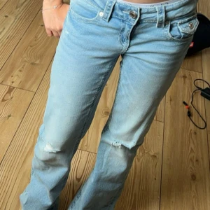 Ljusblå raka jeans med slitningar - Säljer ett par ljusblå jeans med raka ben och snygga slitningar på knäna. Jeansen har klassisk femficksdesign, låg midja och är tillverkade i mjukt denimtyg. Perfekta för en avslappnad och trendig look.