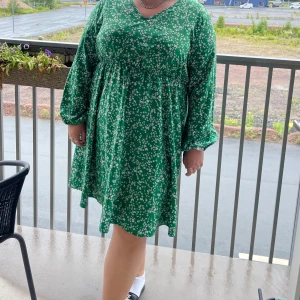 Grön blommig klänning med v-ringning - Superfin grön klänning med små vita blommor, v-ringad hals och långa ärmar. Modellen är kort och har en lös passform som ger en skön och avslappnad känsla. Perfekt för vår och sommar när du vill känna dig både söt och bekväm.