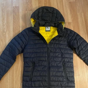 Svart pufferjacka från McKinley - Snygg svart pufferjacka från McKinley med gul insida och huva. Jackan har dragkedja framtill, två sidofickor med dragkedja och quiltad design. Perfekt för kyliga dagar när du vill hålla stilen och värmen.