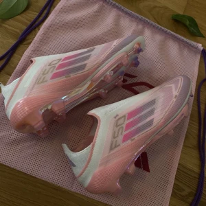 Adidas F50 Elite +, Lamine Yamal par 201 av 304 - Säljer ett par Adidas F50 fotbollsskor i en snygg mix av rosa, lila och vitt. Skorna har Sprintframe 360+ sula för maxad speed och grepp på planen. Ovandelen är i syntetmaterial och designen har coola grafiska detaljer. Perfekta för dig som vill sticka ut på fotbollsplanen.